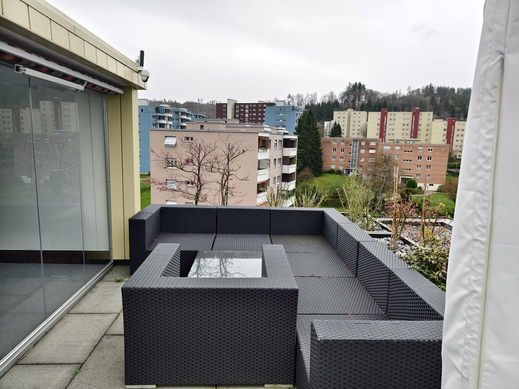 Dachterrasse mit dunkelgrauem Rattan-Sofaset, Blick auf mehrstöckige Wohnblöcke und Bäume.