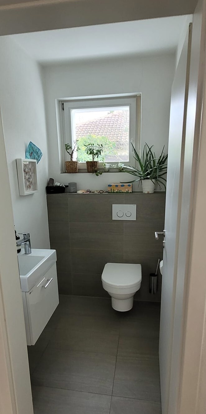 Small bathroom with wall-mounted toilet, vanity sink cabinet on the left, window with plants.