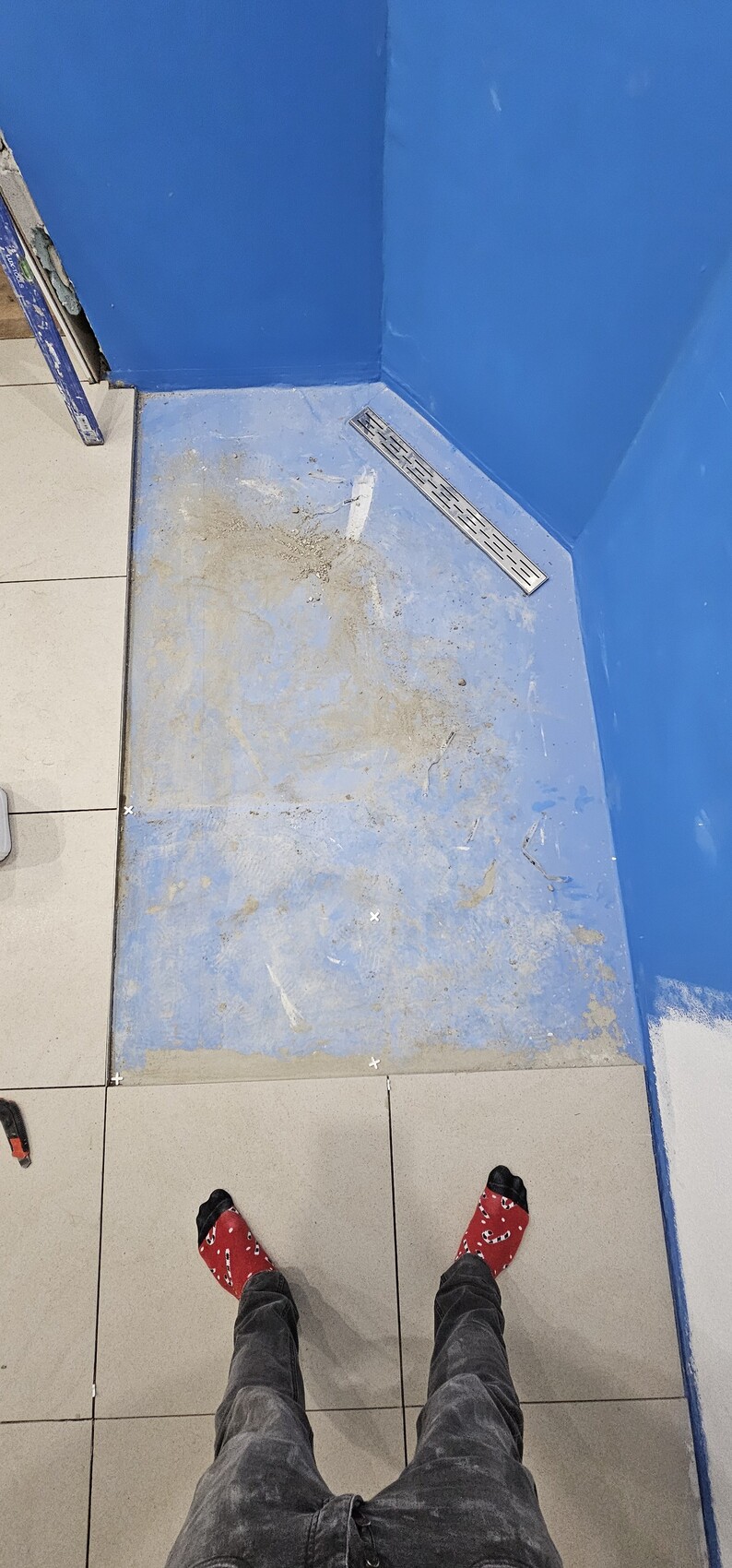 View from above of a blue corner with drain grate and dirty floor; feet in red socks.