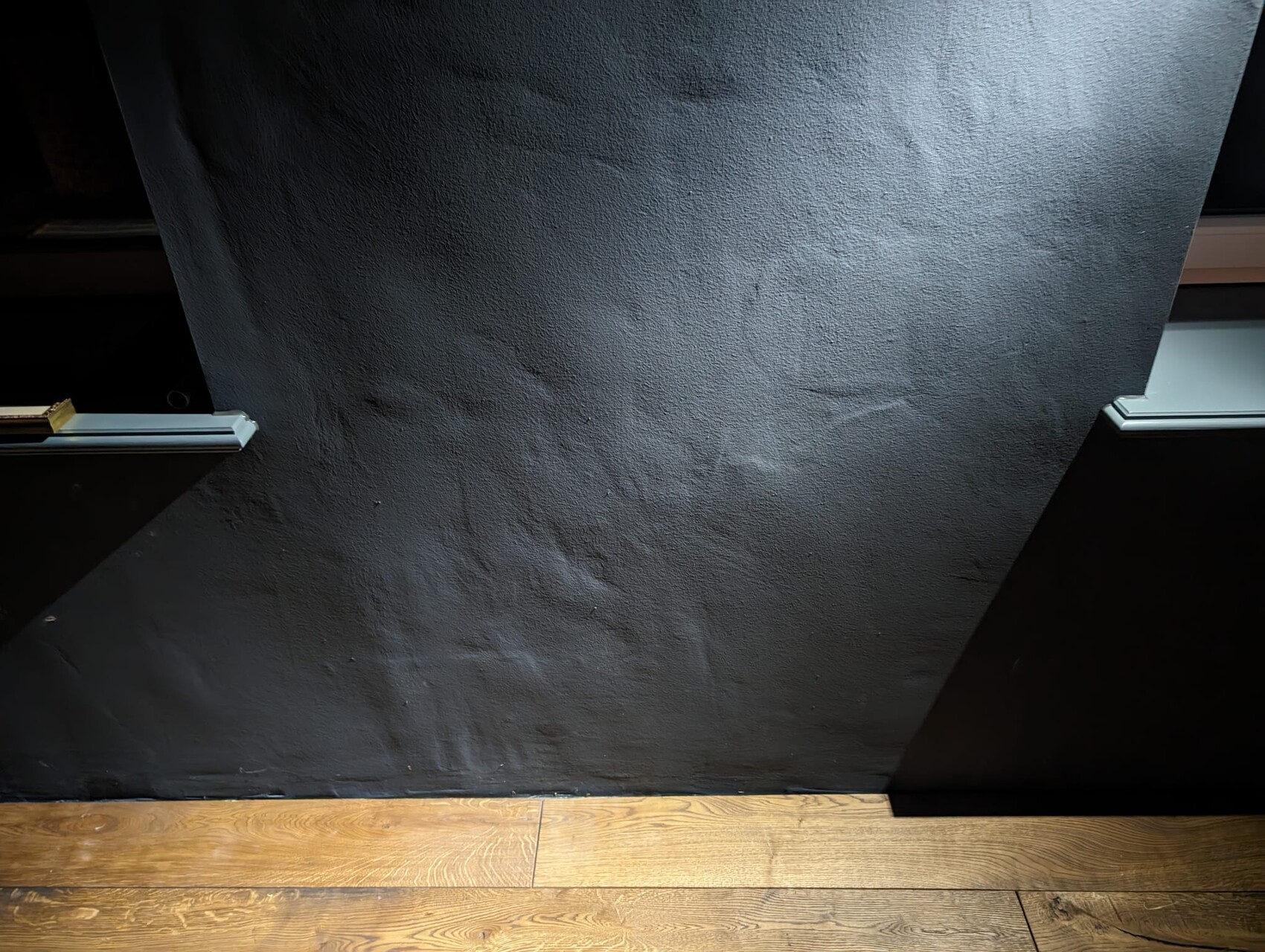 Dark, roughly textured wall above a wooden floor; black panel edges visible at the side.