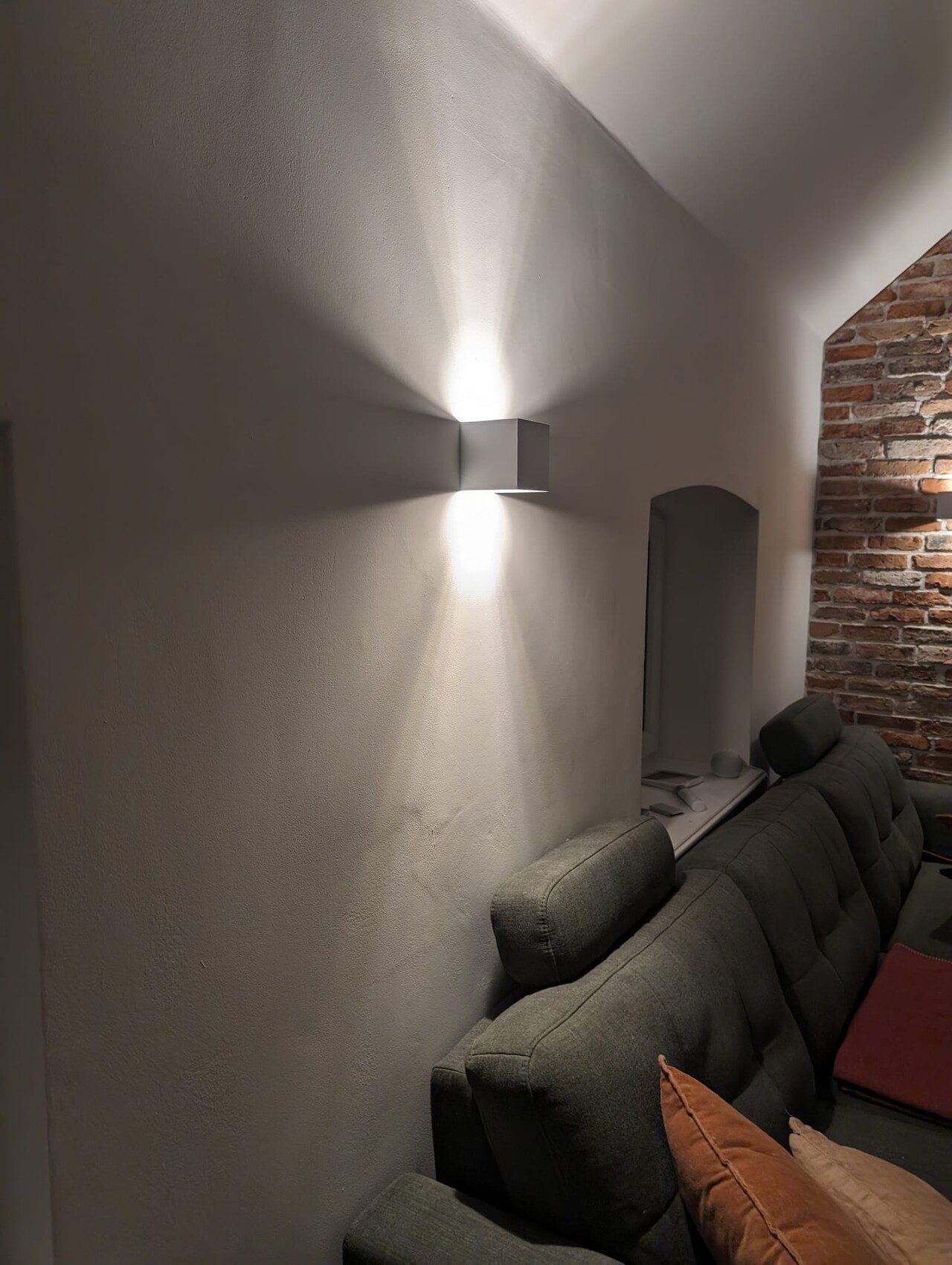 Modern wall lamp on white wall illuminating upwards and downwards; gray sofa, brick wall on the right.