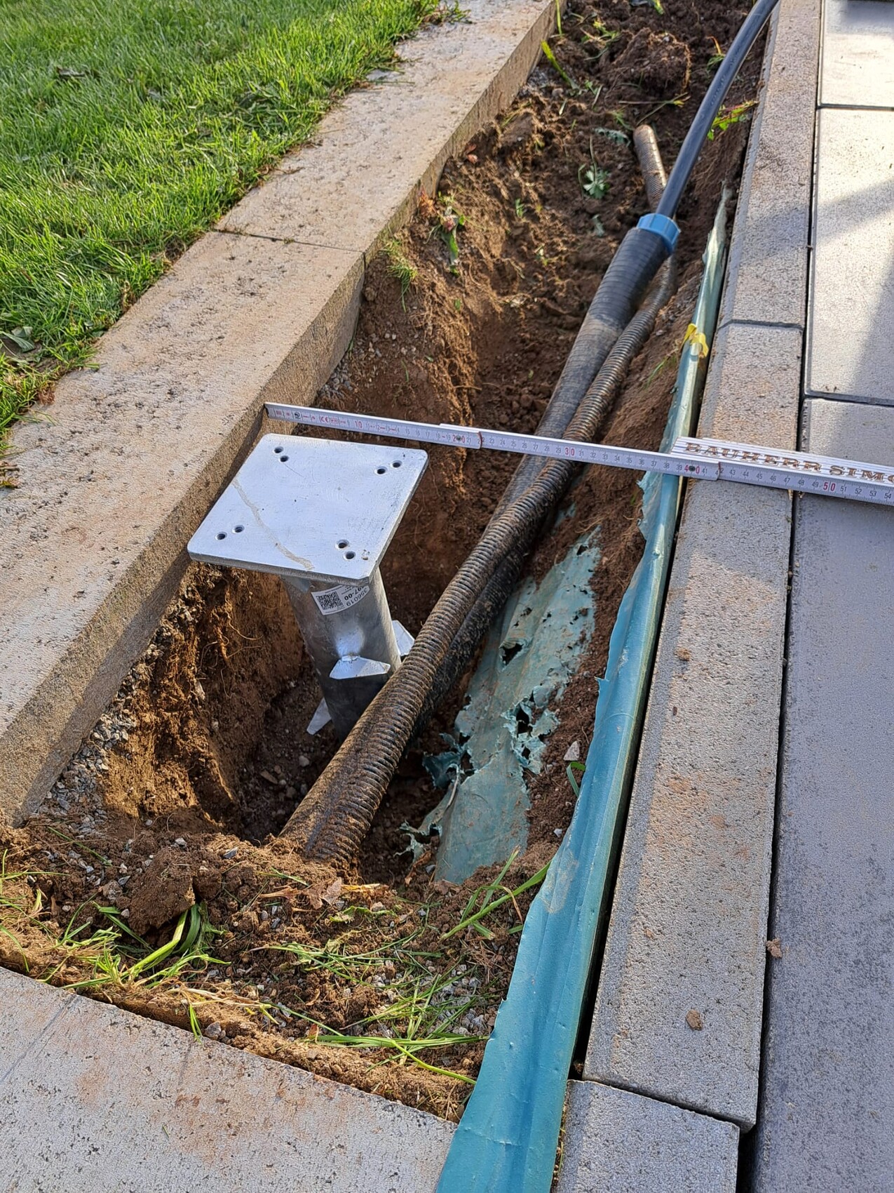 Trench next to sidewalk with pipes, measuring tape and metal base in the soil.