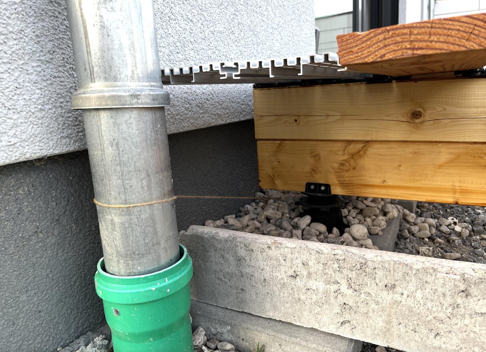 Exterior view: metal downpipe, green pipe connection in gravel, wooden beam substructure.