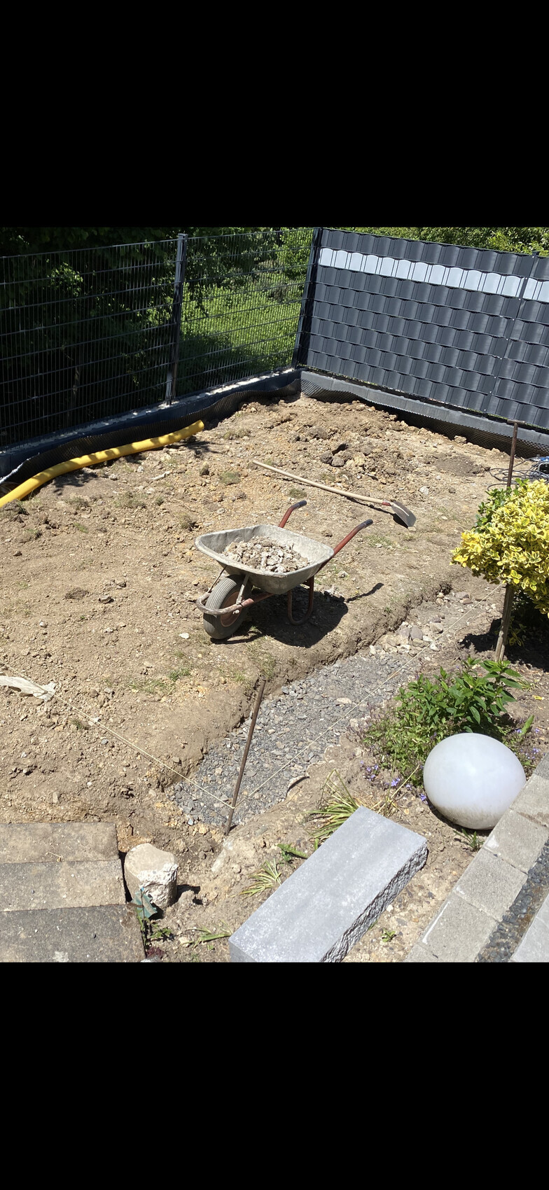 Garden construction site: wheelbarrow with gravel, spade and shovel, loose soil, fence in the background.