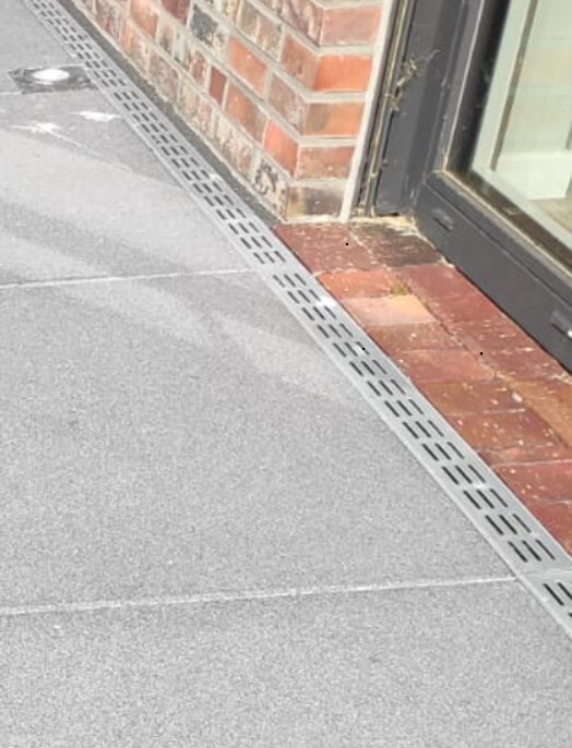 Pathway with metal channel alongside a red brick wall next to a glass facade of a shop.