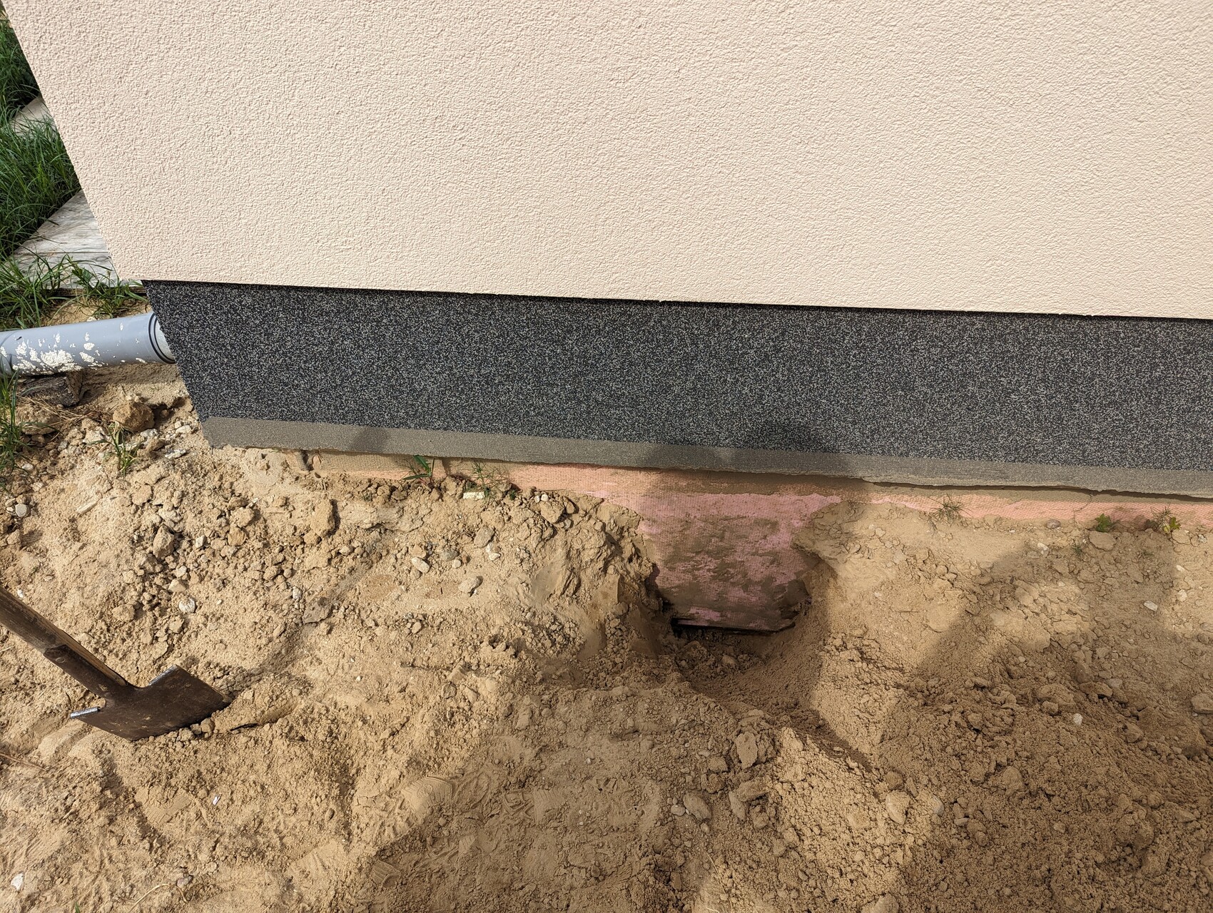 Beige exterior wall with dark base; excavated soil, shovel in front.