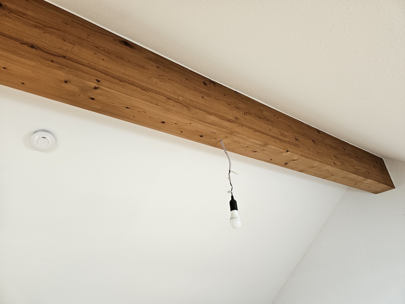 White room with wooden beam; loose cables lead to a hanging light bulb, smoke detector on the wall.