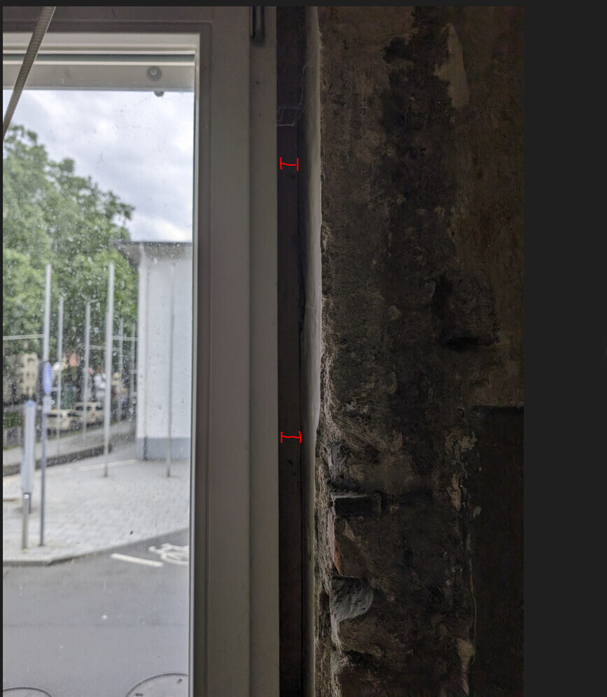 Fensterrahmen neben roher Steinwand; Blick nach draußen auf Straße, Bäume und Gehweg; rote Maßmarken