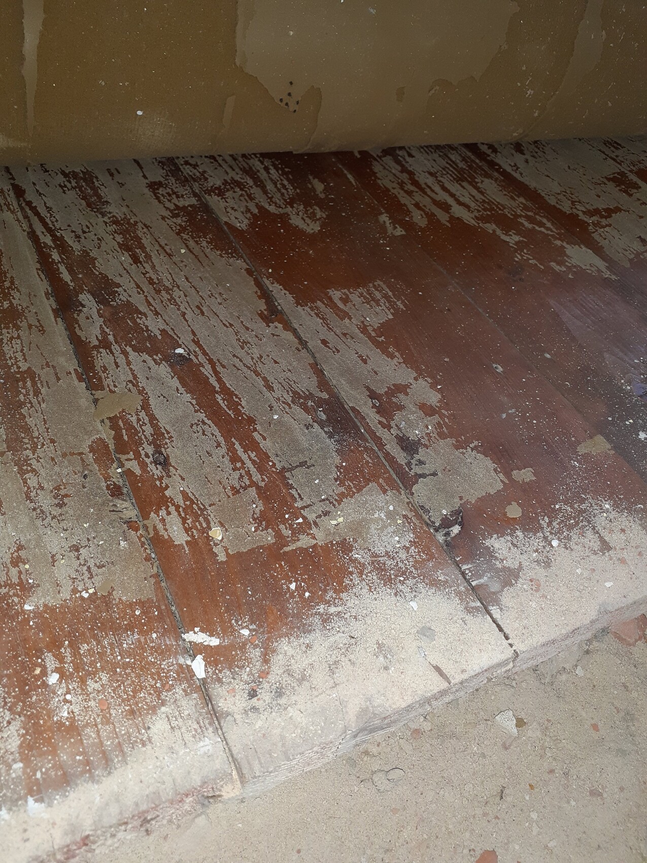 Worn wooden floor with flaking varnish, dust and dirt; wall with plaster peeling off.