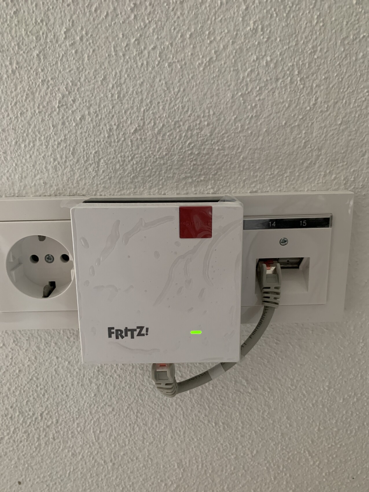 White FRITZ! router plugged into wall socket, green LED lit, network cable connected.