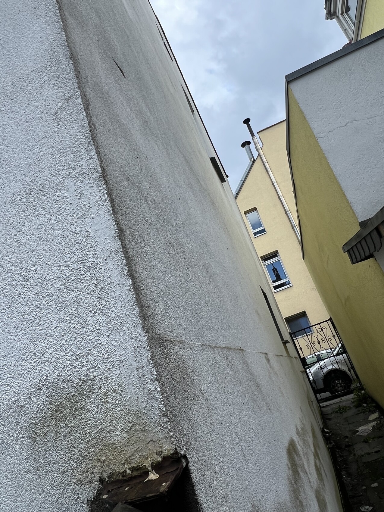 Enge Gasse zwischen weißen Wänden und gelbem Haus, Tor mit geparktem Auto am Ende.