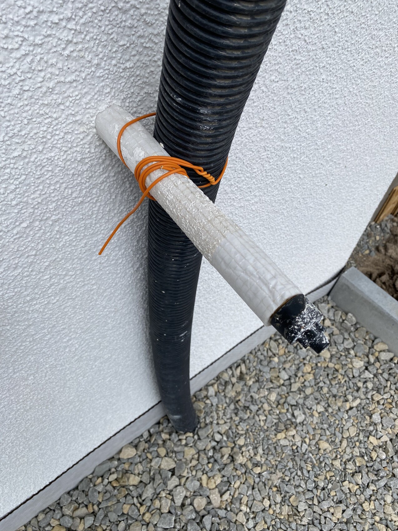 Aussenwand mit schwarzen Rohrleitungen, weisses Isolationsrohr und orange Kabel, Kiesboden.