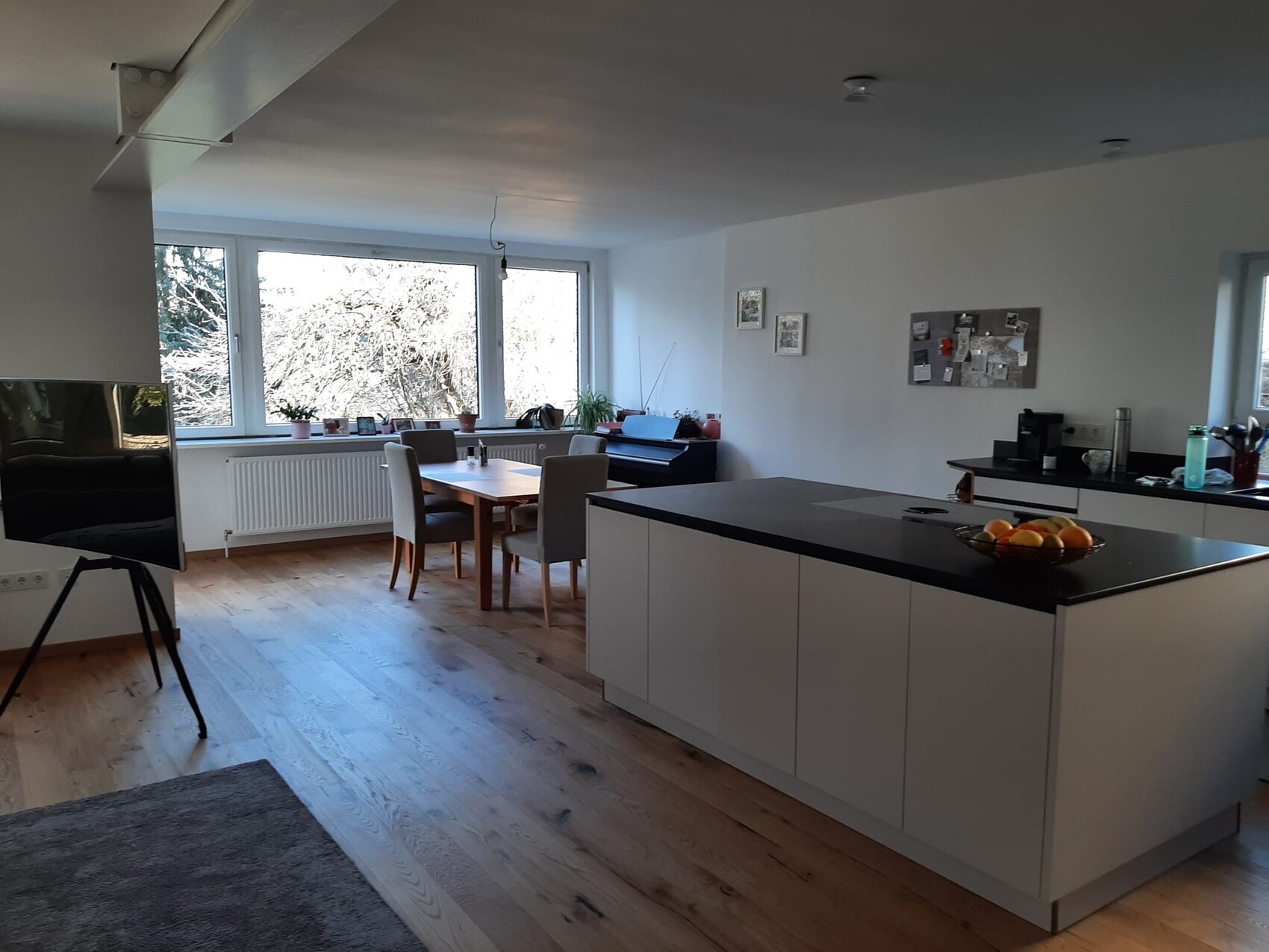 Bright open kitchen area with island, dining table, large windows, and a bowl of fruit.