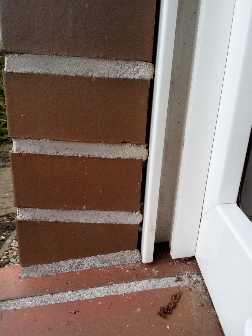 Close-up of a red brick wall next to a white window frame; dusty ground with gravel.