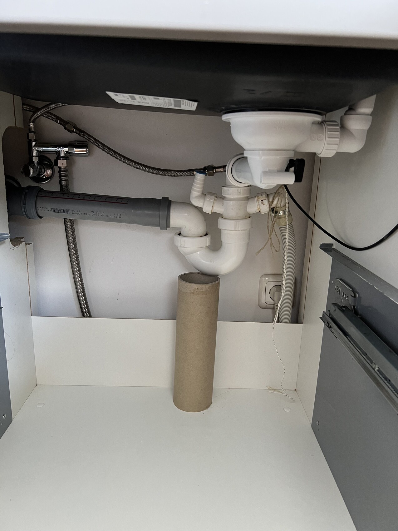 Under the kitchen sink visible drain pipes (P-trap) and a cardboard pipe on the floor.