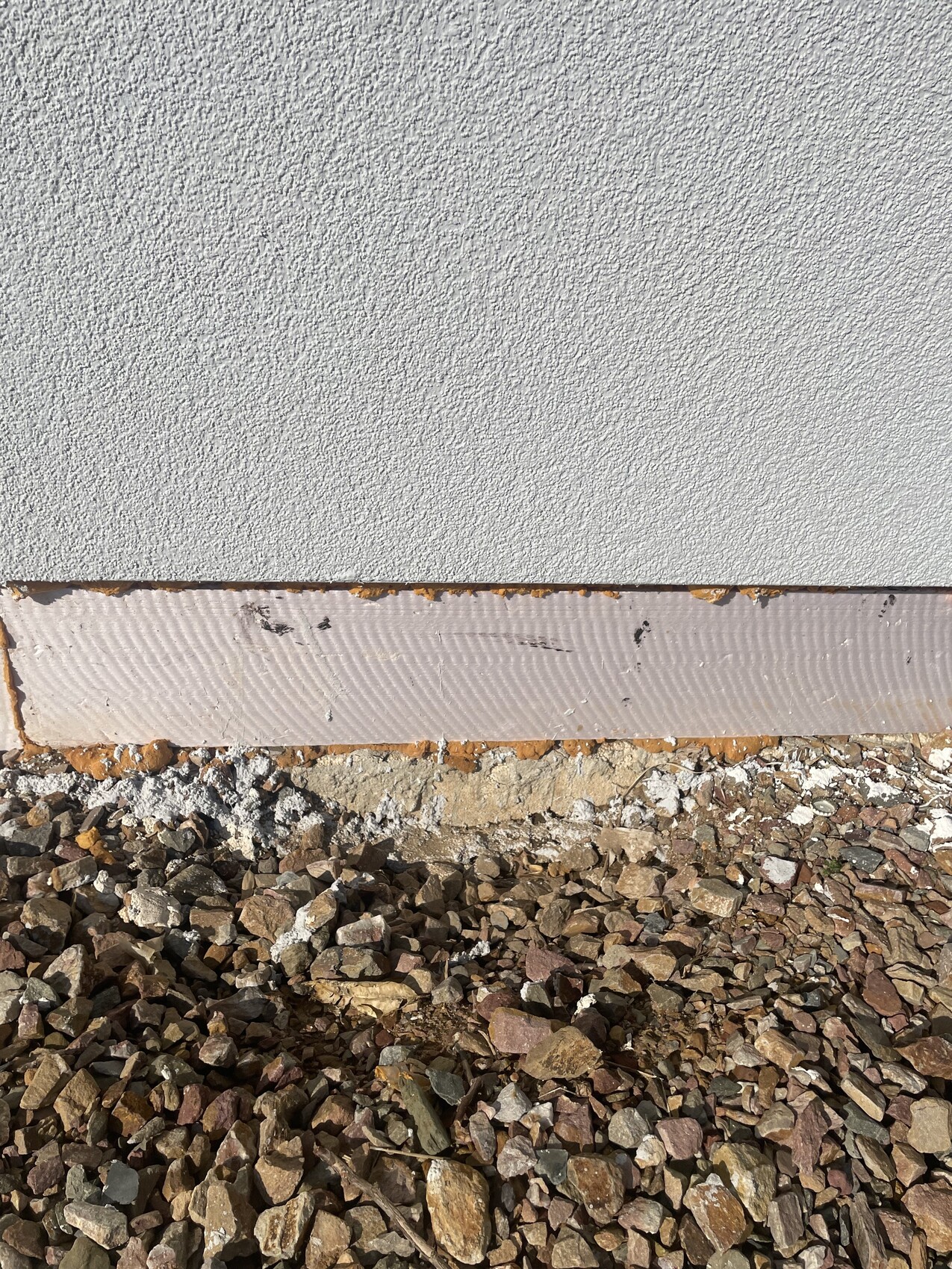 Exterior wall with gray plaster, below it a pink insulation layer and gravel base.