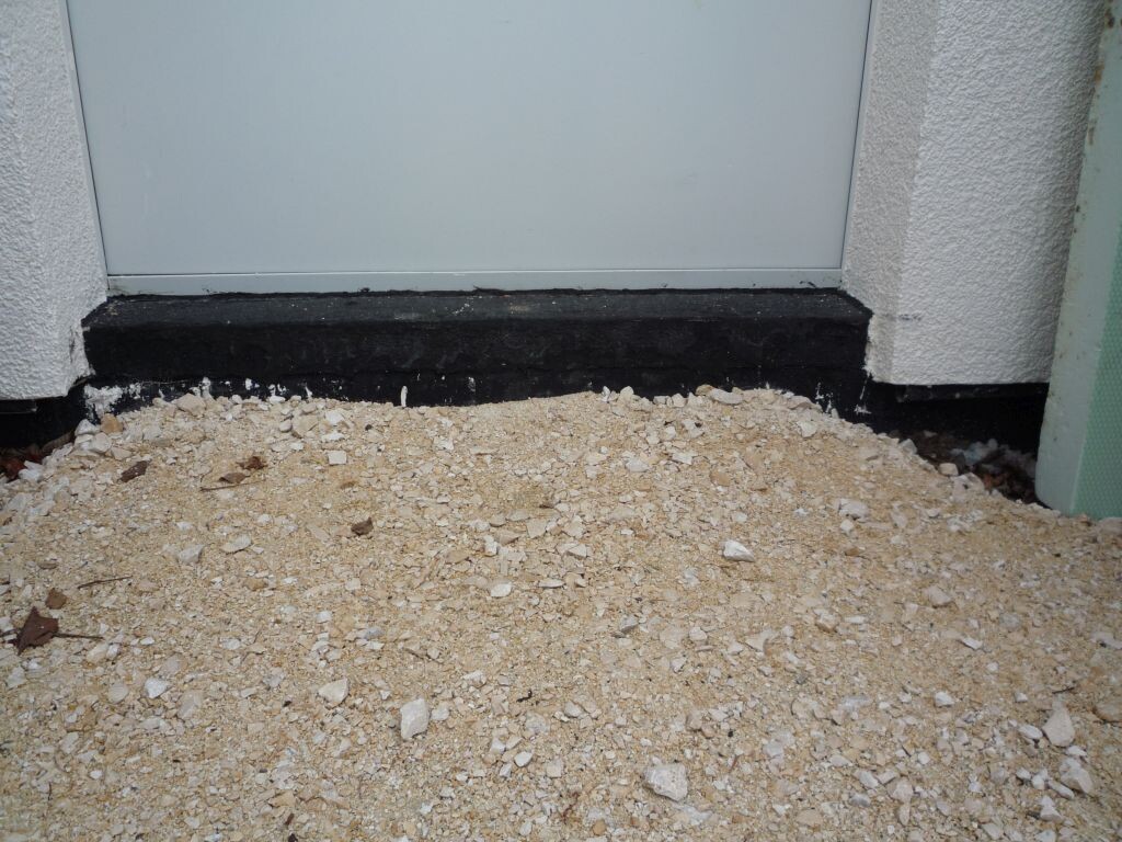 Exterior area in front of a front door with gravel floor, black sealing strip, and white facade.