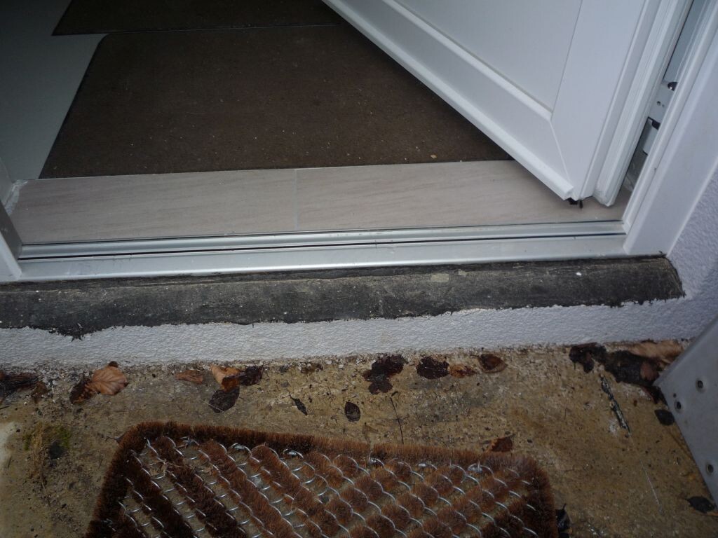 Open entrance door with threshold, interior floor visible, outside a brown doormat and autumn leaves.