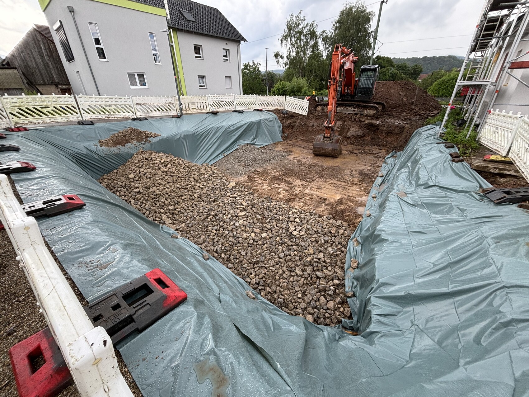 Excavation covered with blue plastic sheeting, gravel piles and excavator next to houses.