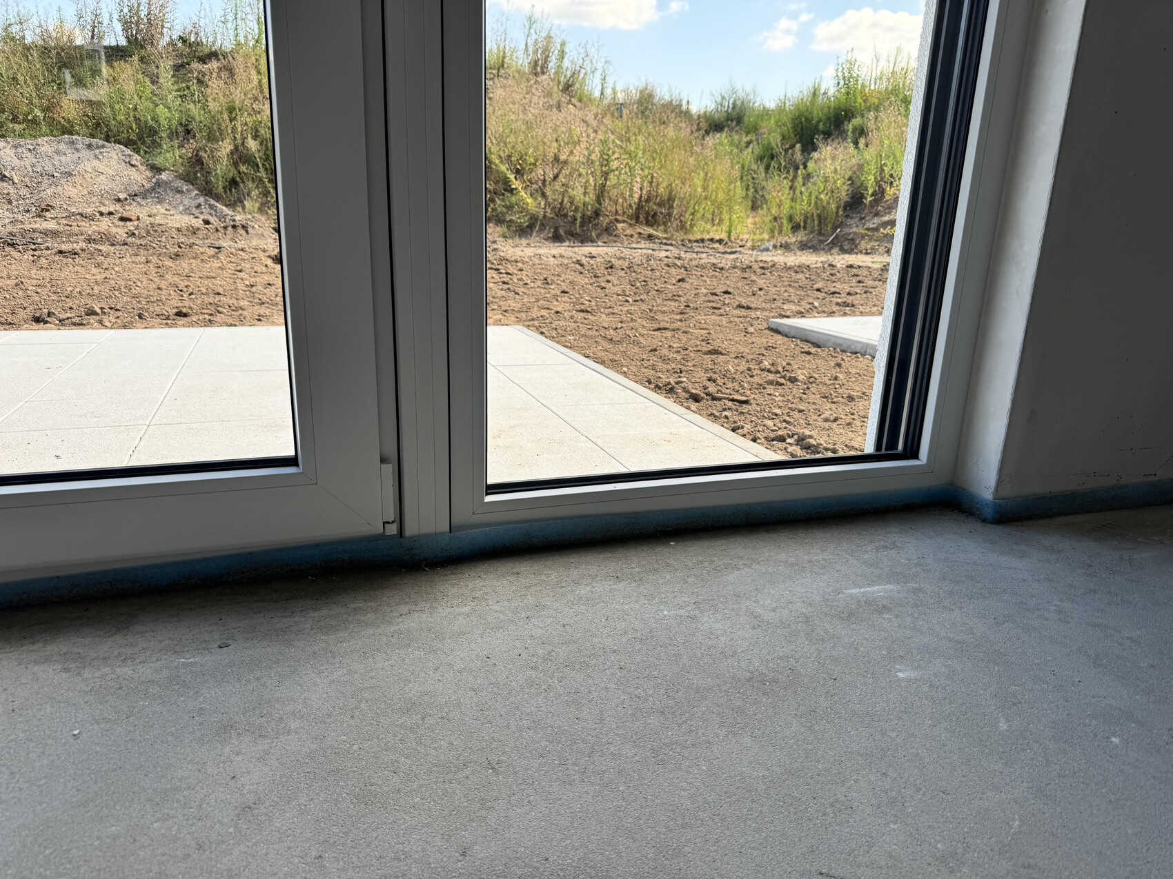 View through white glass sliding door onto bare soil and concrete path, interior with concrete floor.