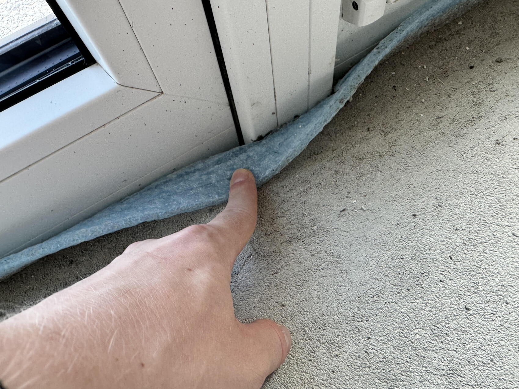 A finger touching a blue insulation roll at the window frame above raw concrete floor.