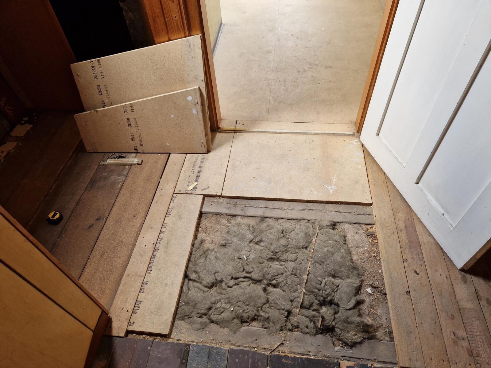Open floor area during renovation showing exposed insulation, wooden planks, and door frame.