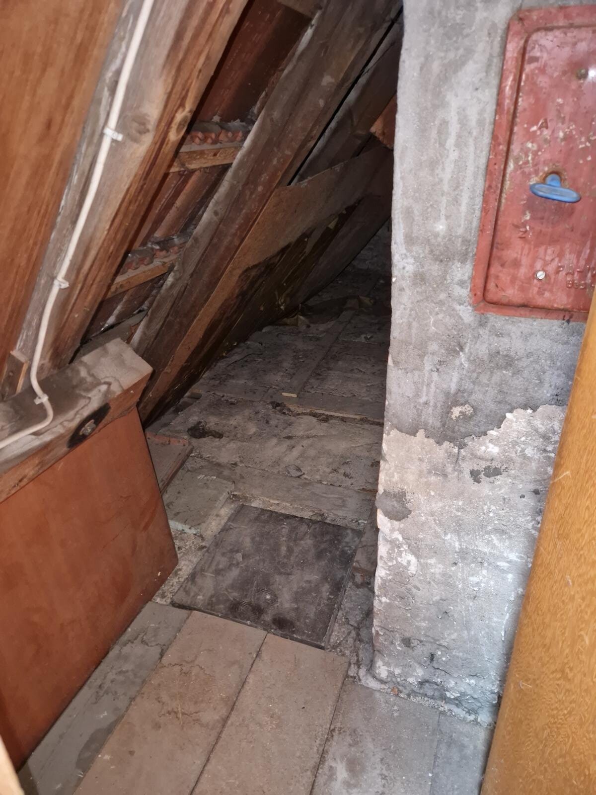 Attic with sloping wooden joists, dusty floor, concrete support, and red wall panel with blue handle.