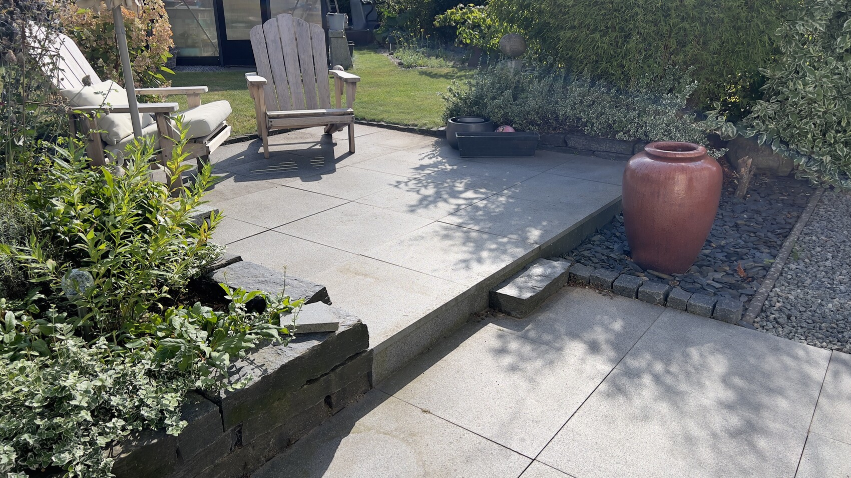 Terrassenbereich mit zwei Holz-Stühlen, roter Keramikvase, Topfpflanzen und Stufen an der Terrasse.