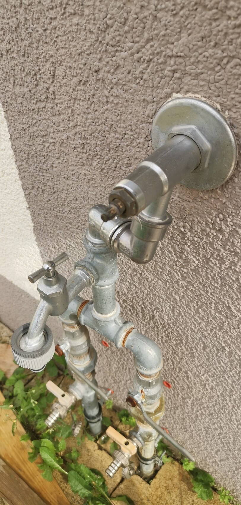 Outdoor faucet with several valves and piping on a rough wall, garden soil.