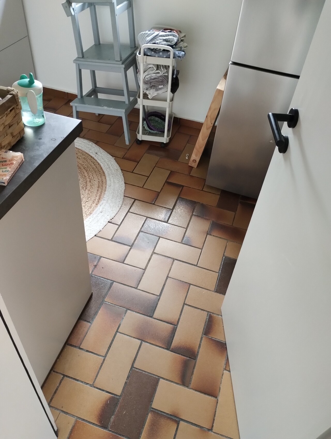 Small utility room: brown floor tiles, refrigerator on the right, shelves and laundry trolley with towels.