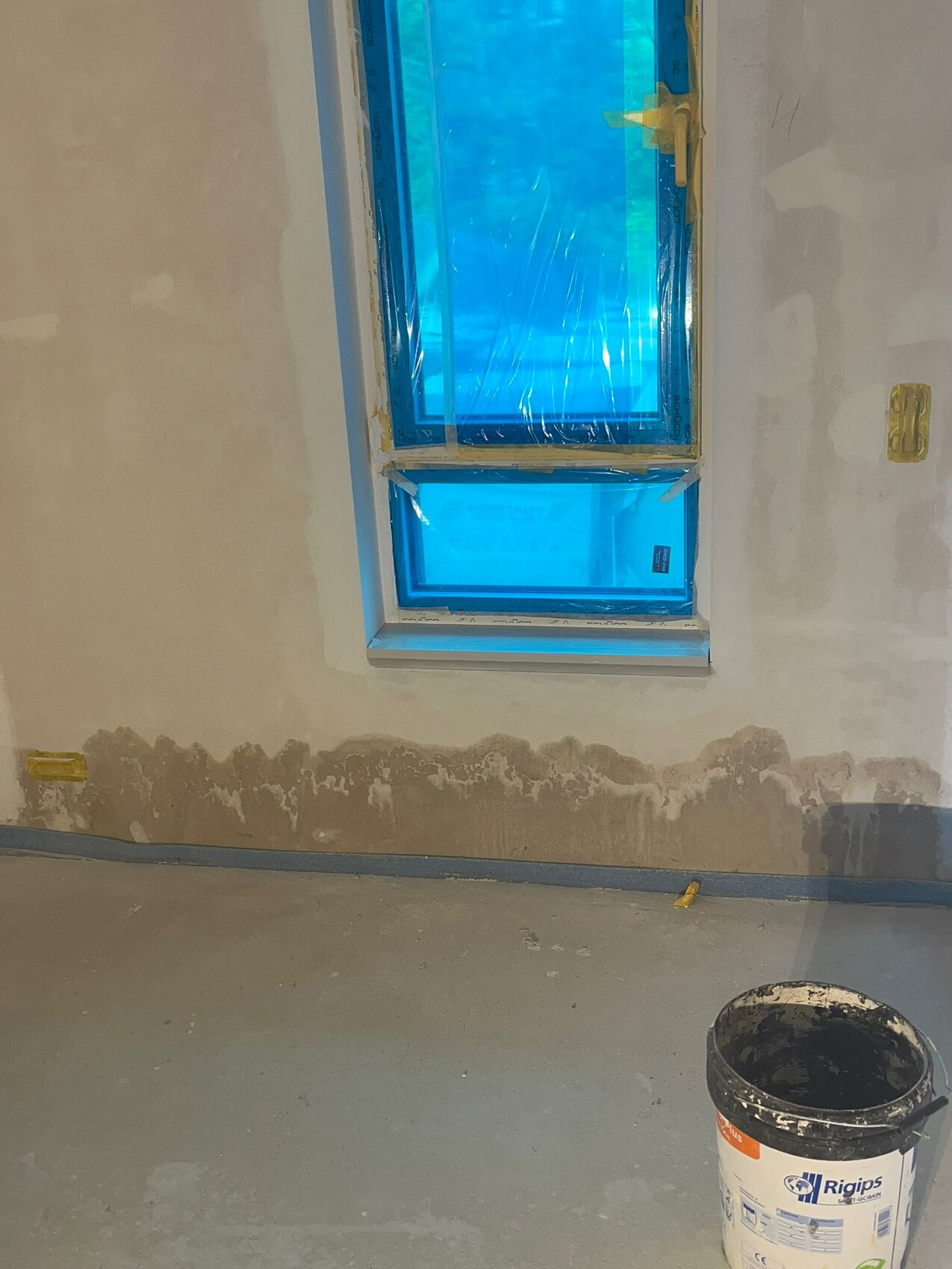 Renovation room with window covered in blue tarp, unfinished walls, and bucket on the floor.