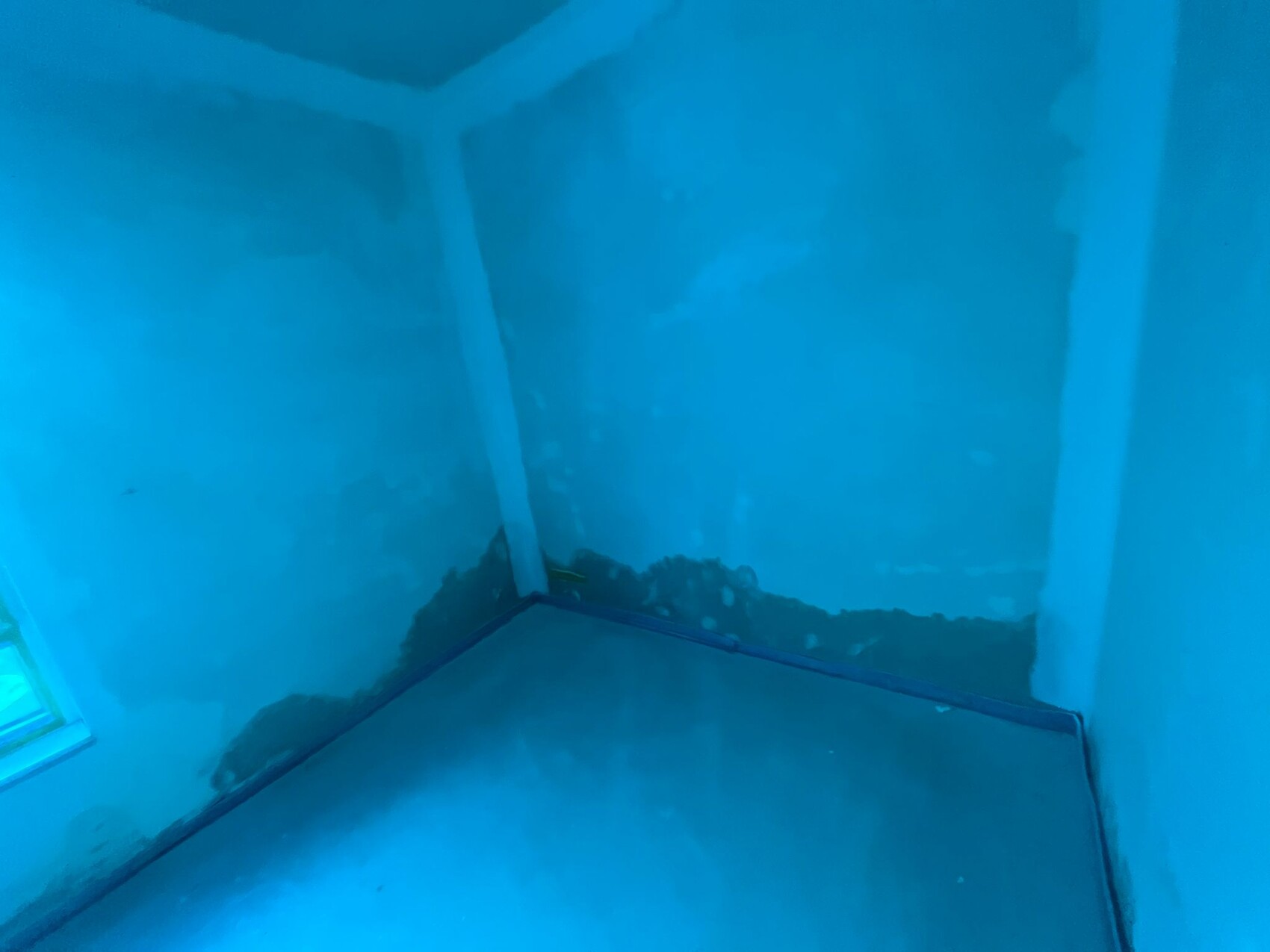 Corner of an unfinished room illuminated by blue light with damp plaster spots on the walls.