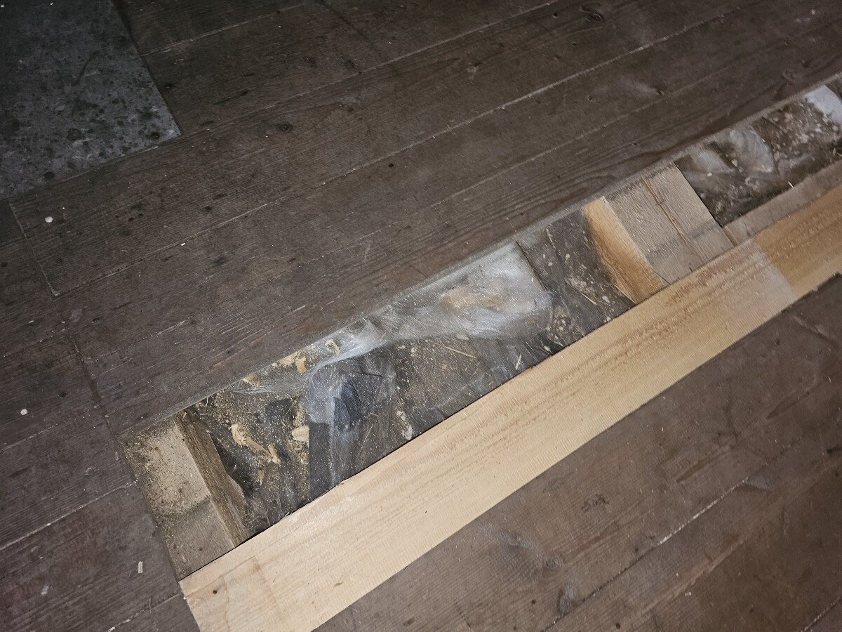 Open floor gap with wooden beams, dust, and visible insulation material.