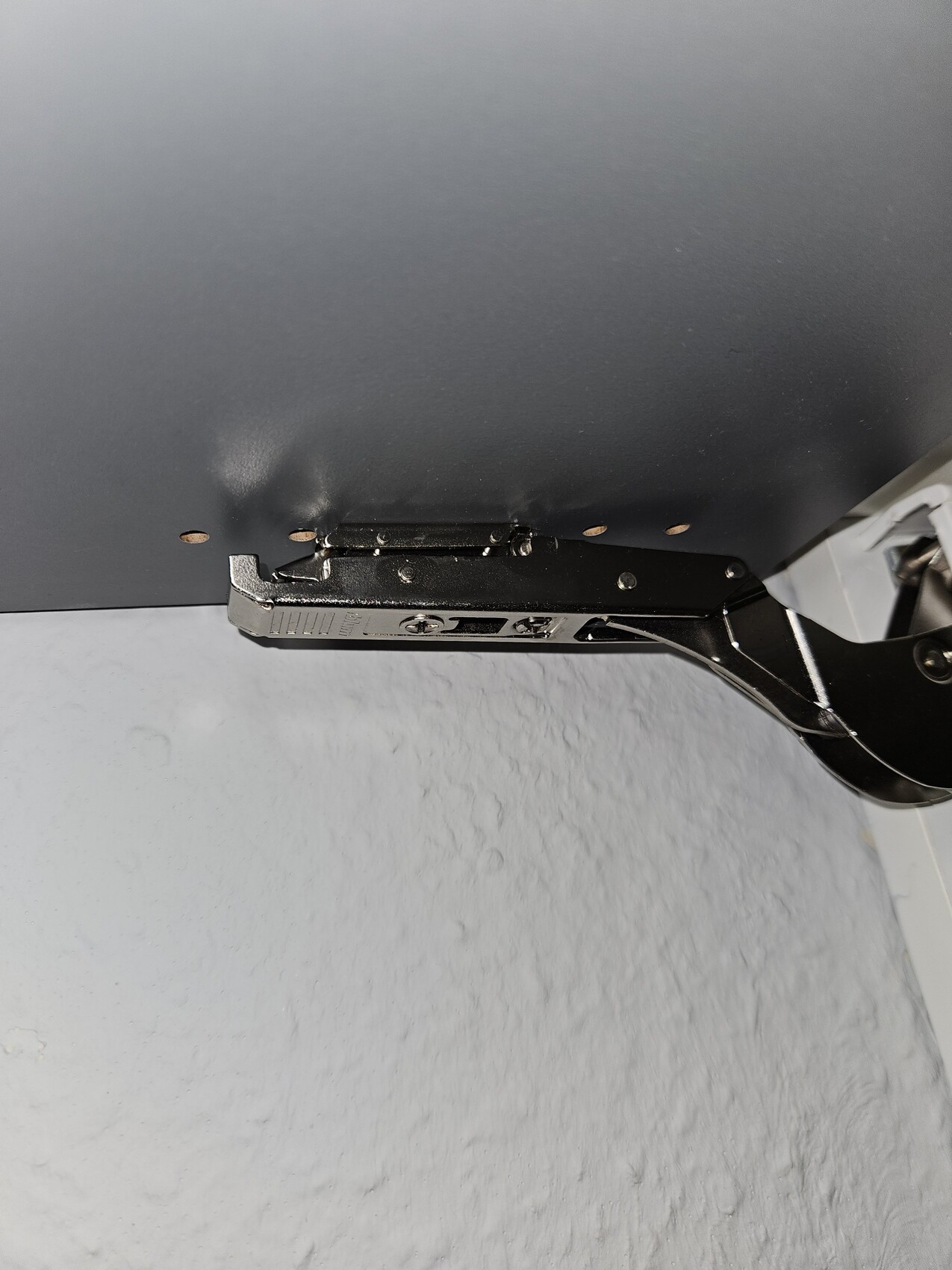 Underside of a grey panel with black metal hinge arm and screws