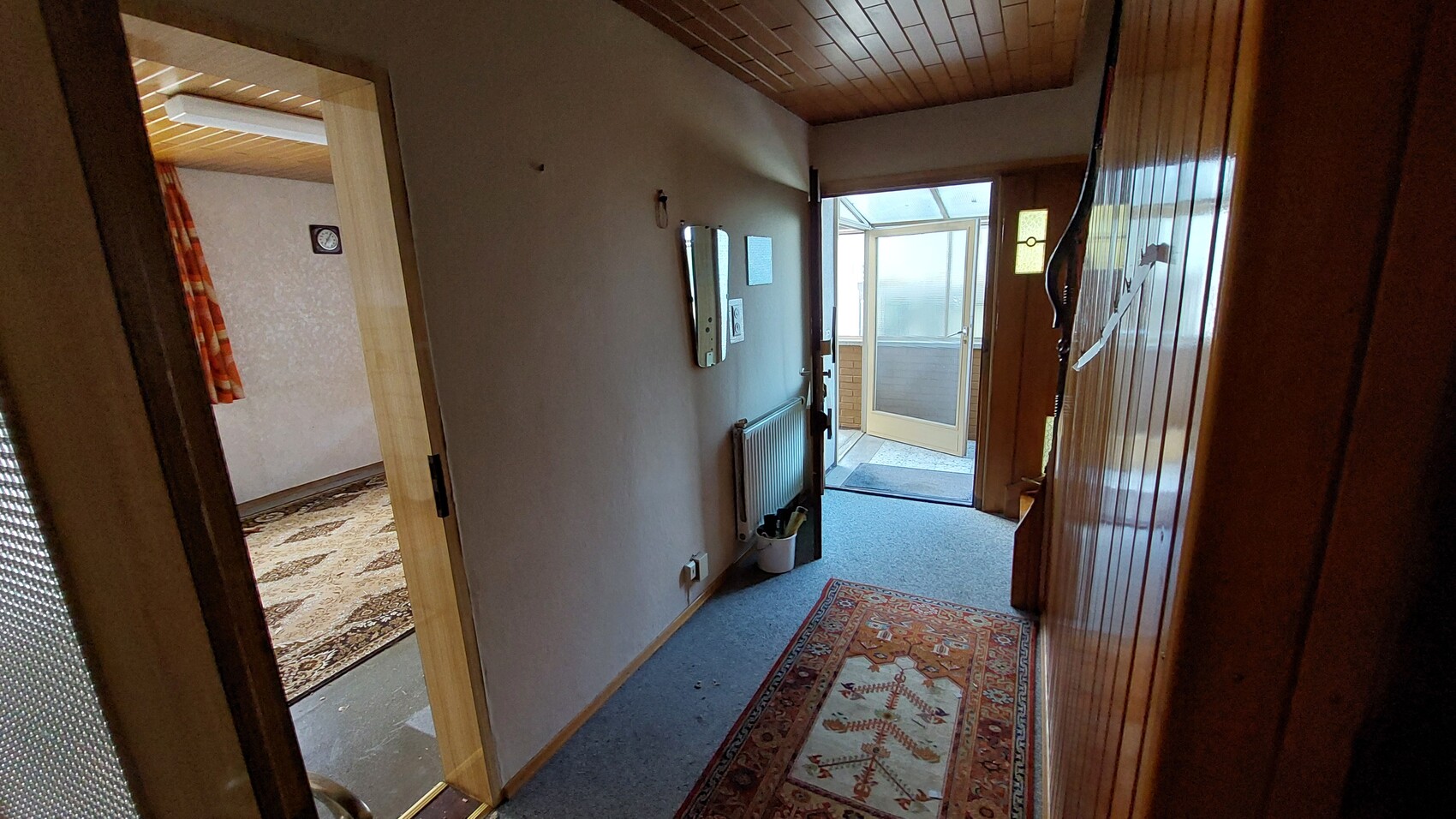 Bright hallway with wooden floor and ceiling, left room with patterned carpet and curtain, glass door at the end.