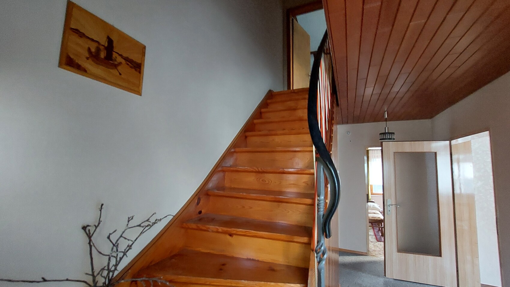 Wooden staircase in hallway with railing, open door to bedroom, wall art on left, dry branches on floor.