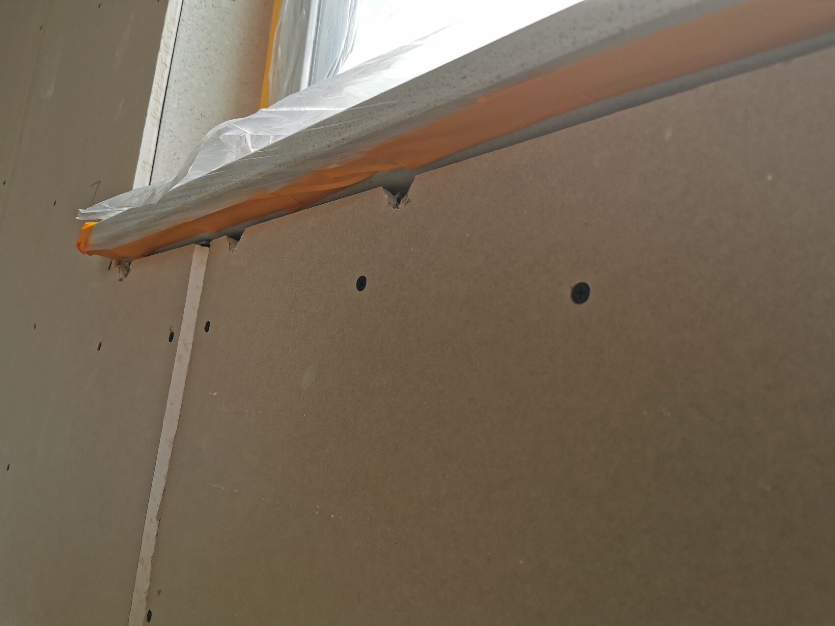 Close-up of drywall panel with screws, top edge protected by film and orange tape.