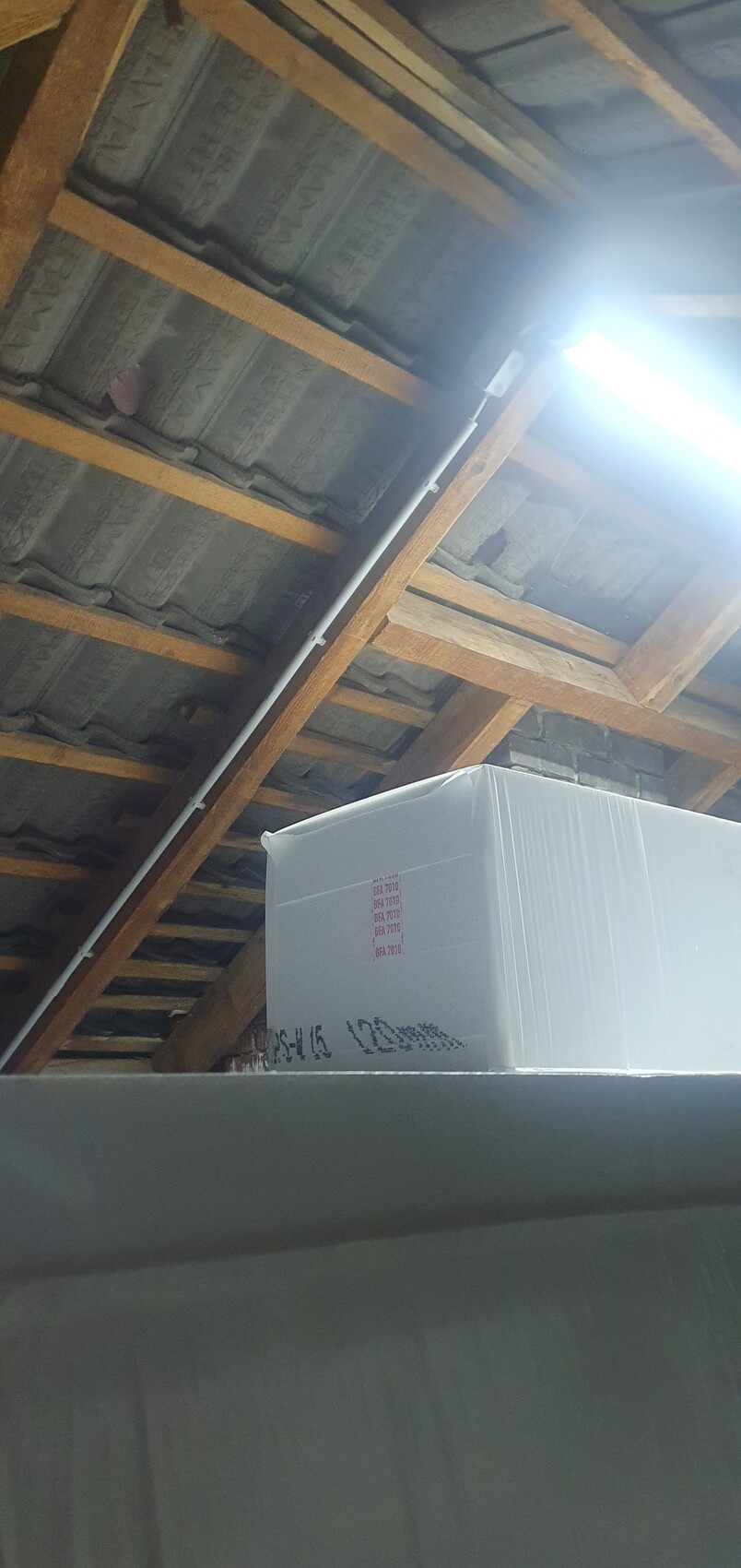 Dachboden mit Holzbalken; großer, weiß verpackter Karton und helles Licht von der Decke