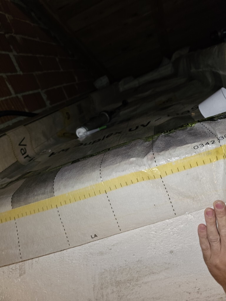 Attic construction site: yellow tape measure over insulation membrane, right hand at edge, brick wall in the background
