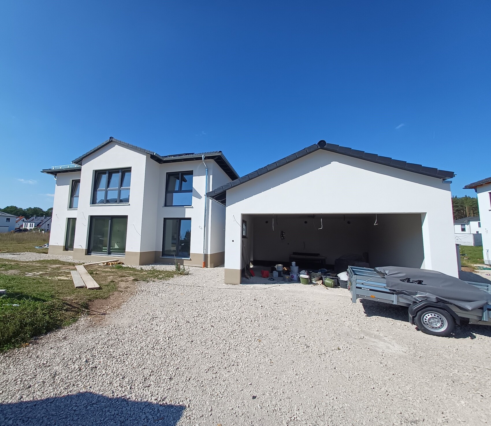 Modern white detached house with open garage, construction tools and trailer in front.