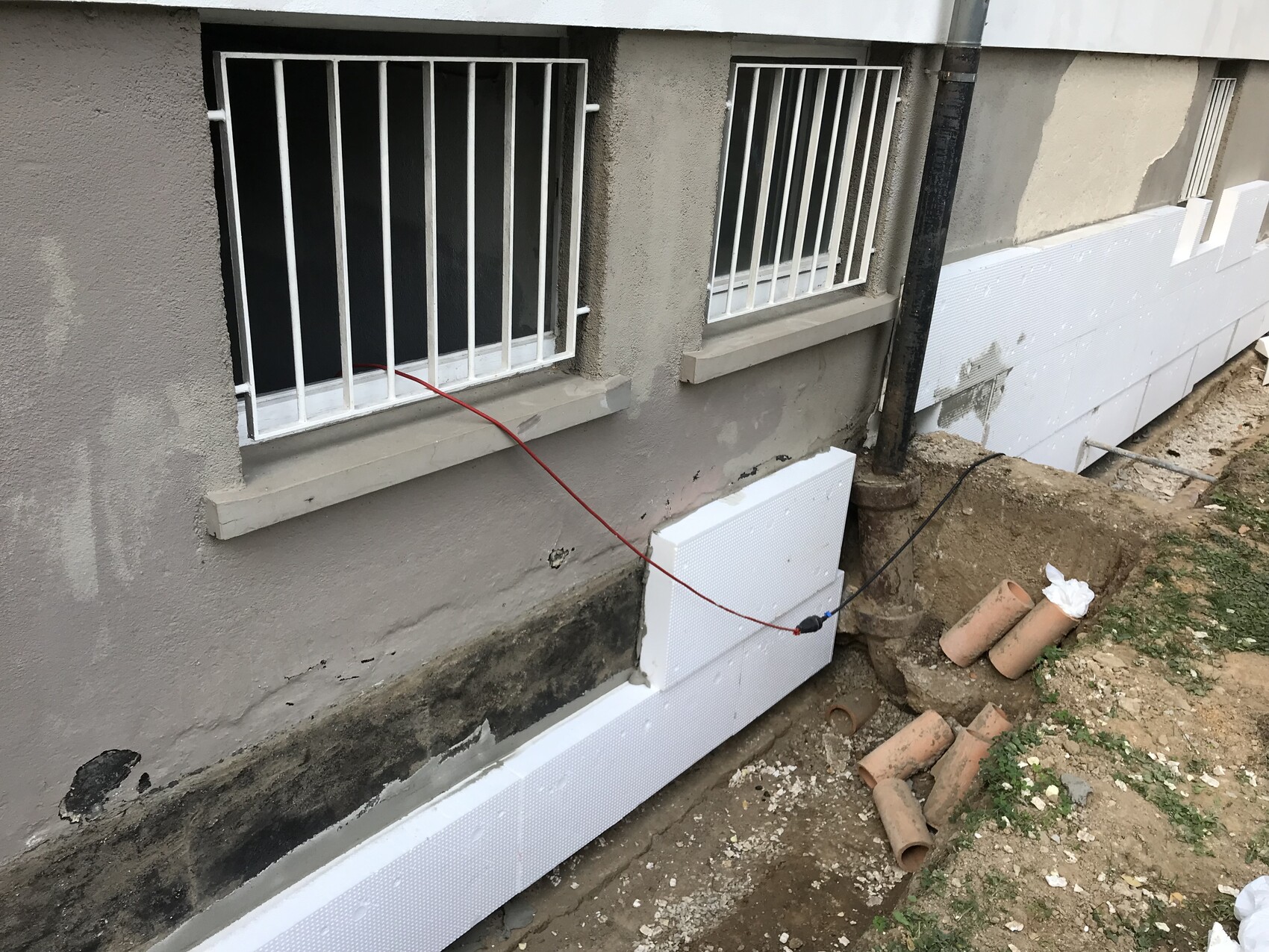 Außenwand mit Gitterfenstern, am Fundament Dämmplatten montiert, Bauabfall und Rohre.