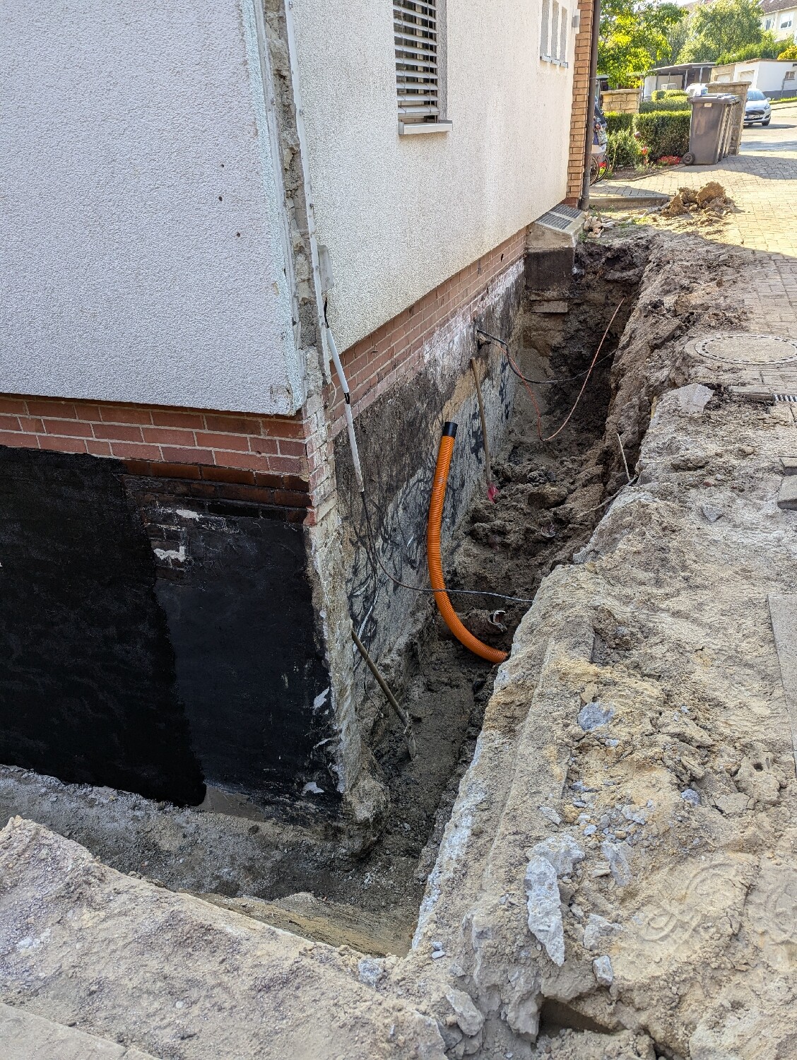 Deep excavation next to the house foundation with orange hose and earthworks in the yard.