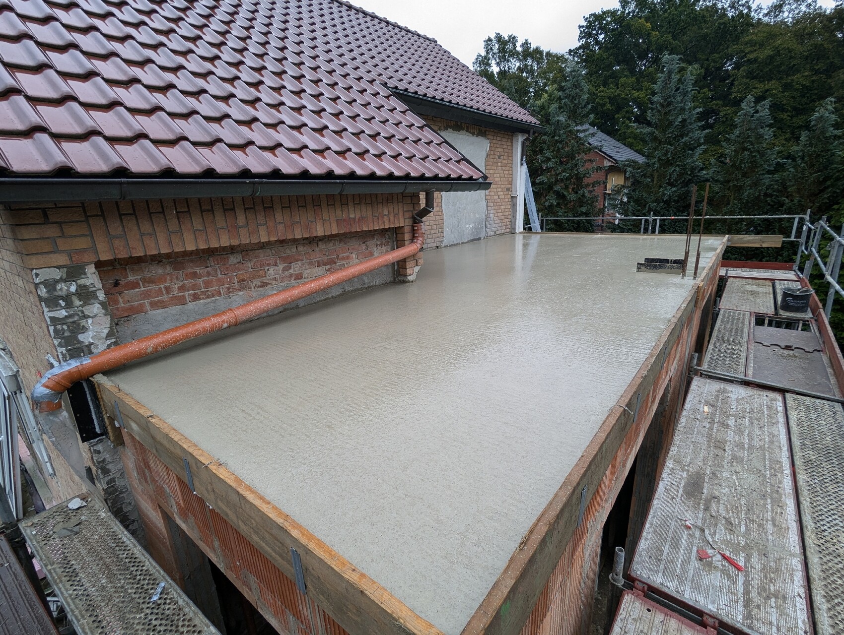 New concrete floor slab on extension; brick wall, red tiled roof, orange downpipe, scaffolding.