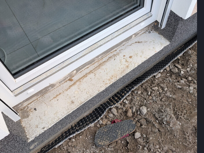 Window sill with concrete frame, in front soil and gravel, black drainage strip on the ground.