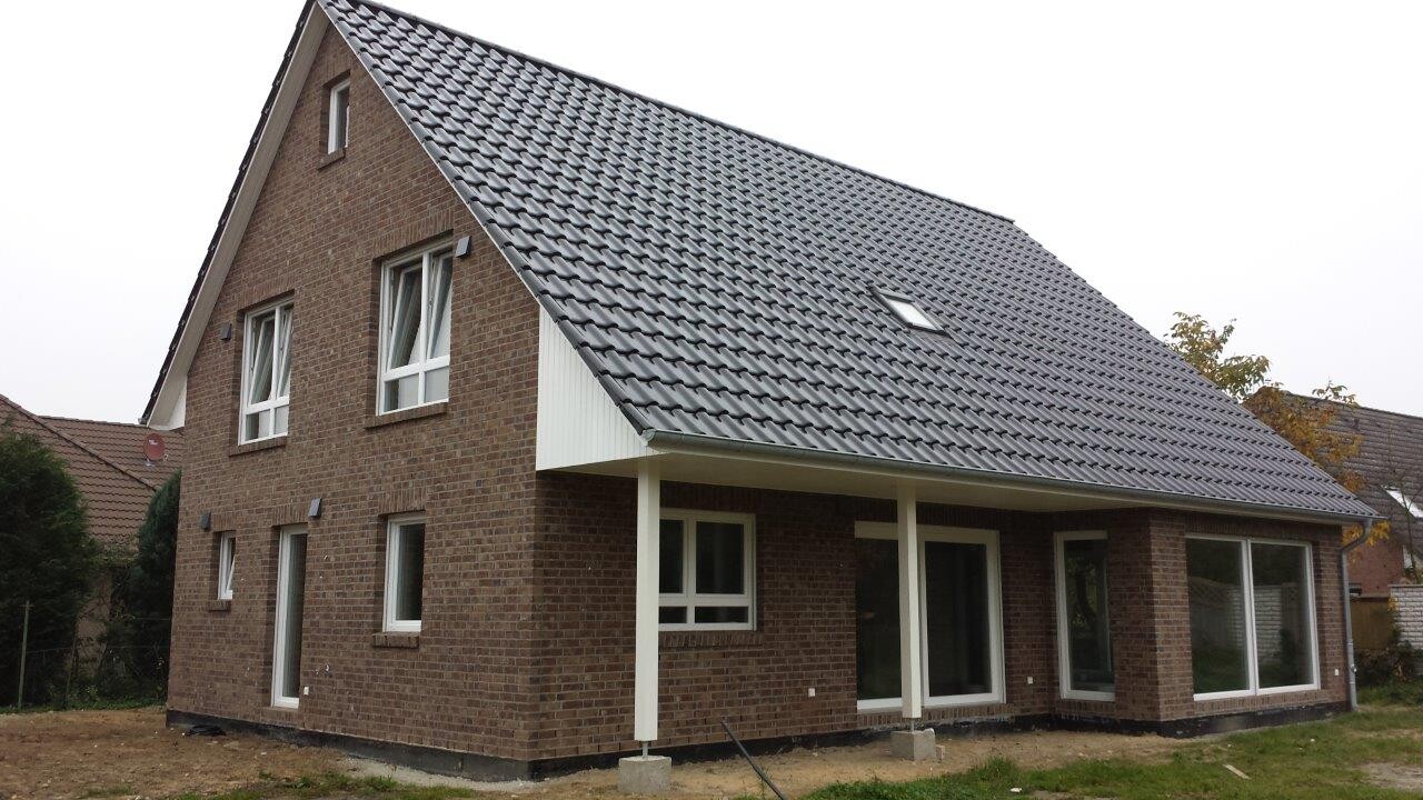 Modern brown brick house with steep gray tiled roof, large windows, and veranda.