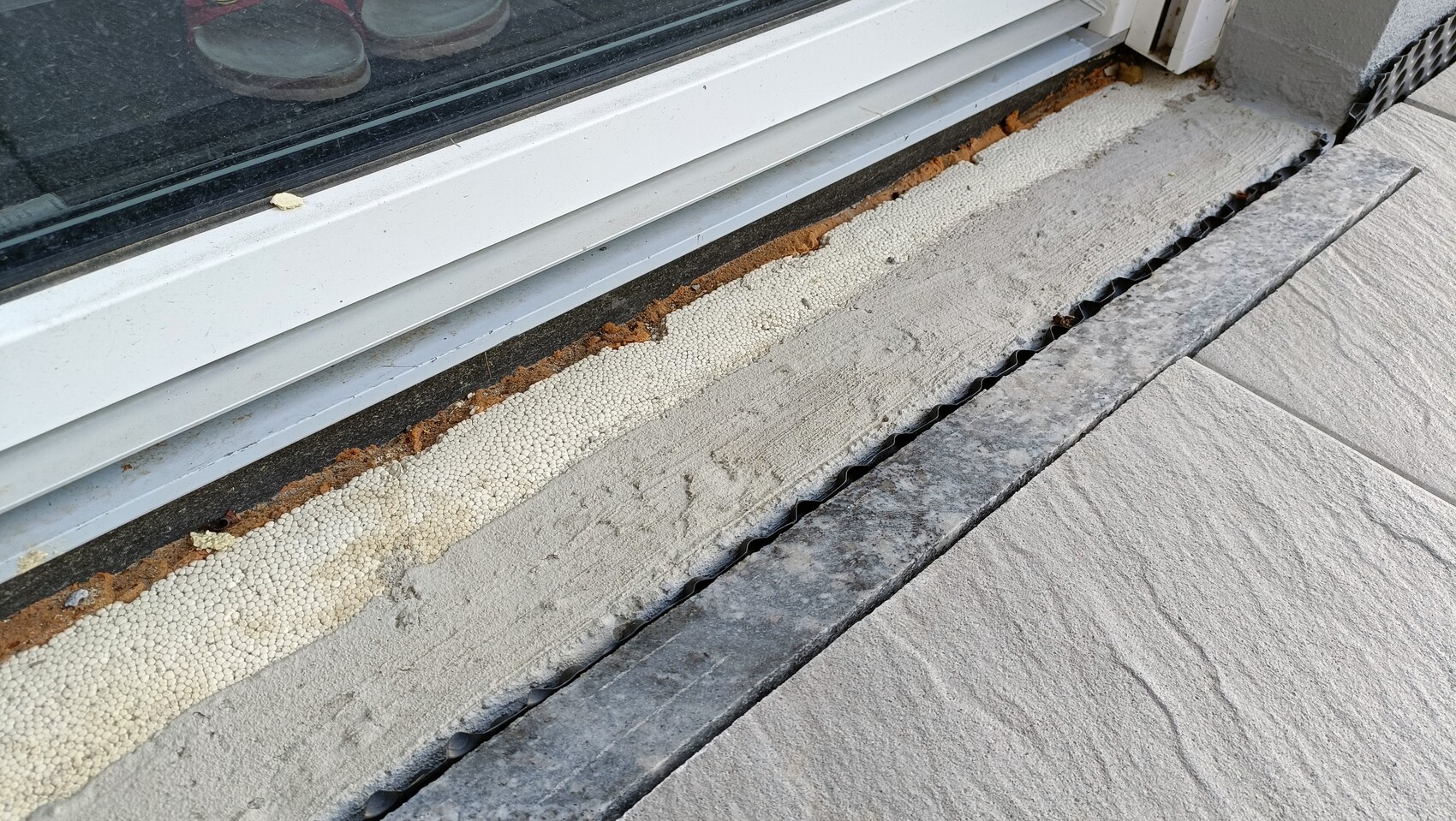 Exterior view: door frame with seal, white foam joint filler, rust, stone slabs, and drainage grate