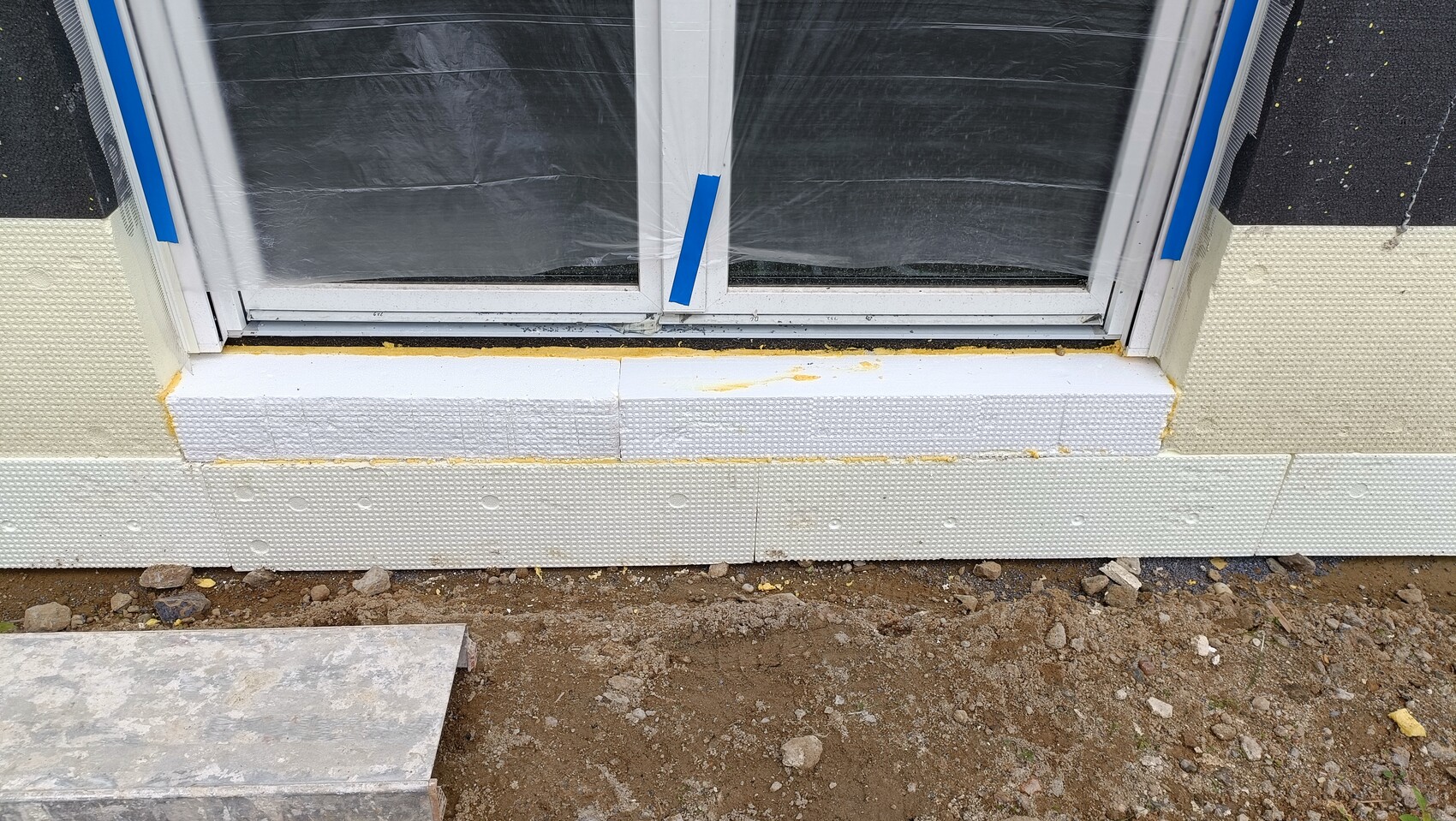Window frame during construction phase, surrounded by white polystyrene boards; blue tape on the frame.