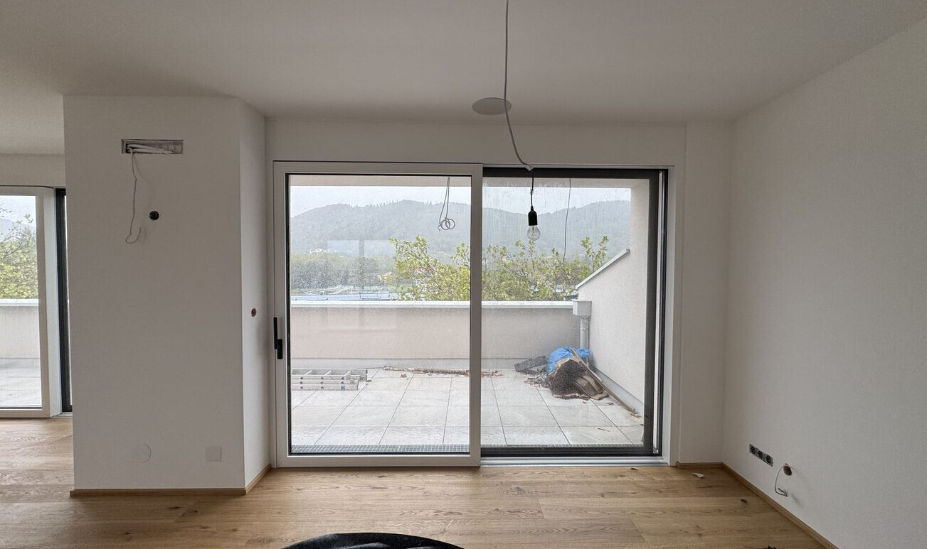 Living room during construction with wooden floor, exposed cable, glass sliding door to the terrace, and mountain view.