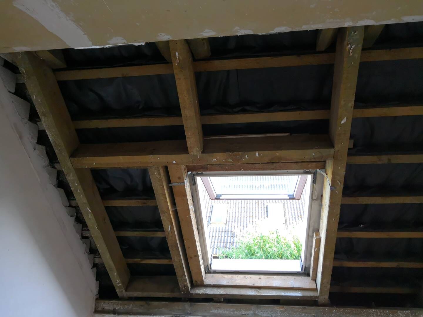 Attic with wooden beams, dark insulation, and open roof window looking outside.