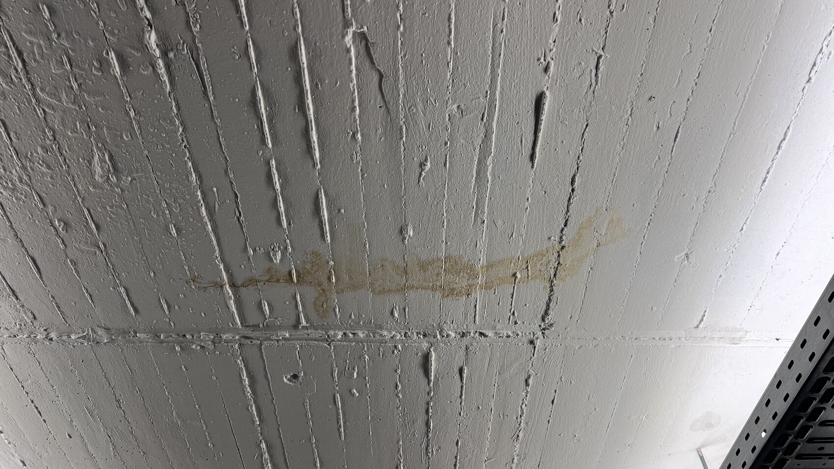 Weiße Deckenplatte mit vertikalen Riefen, braune Flecken entlang einer Linie; rechts unten Metallregal-Kante.